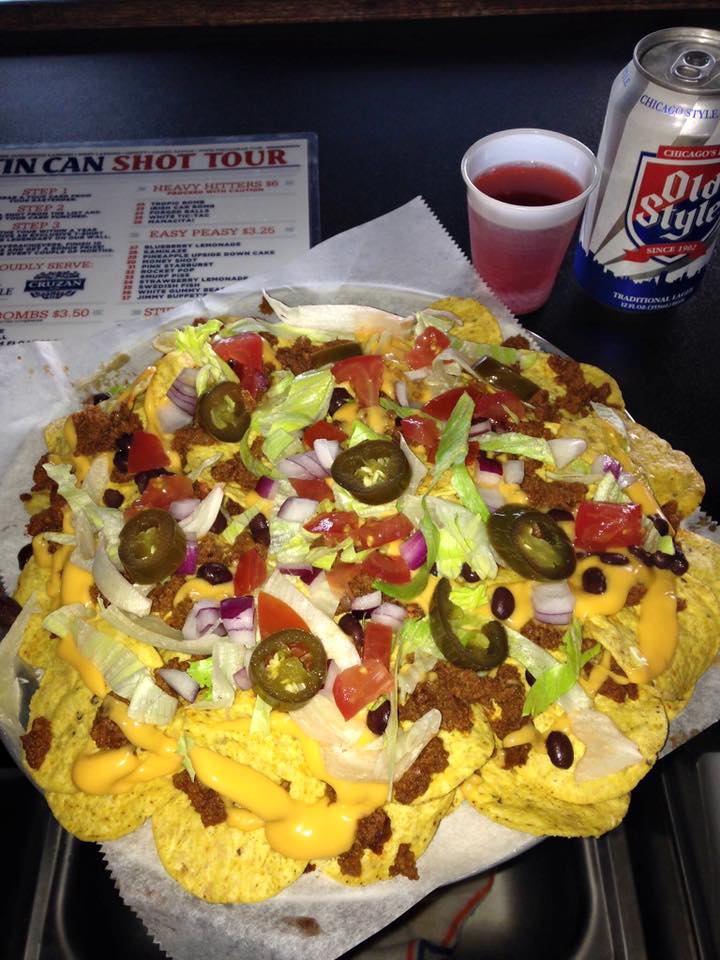A plate of loaded nachos with a can next to a Tin Can Shot Tour menu.